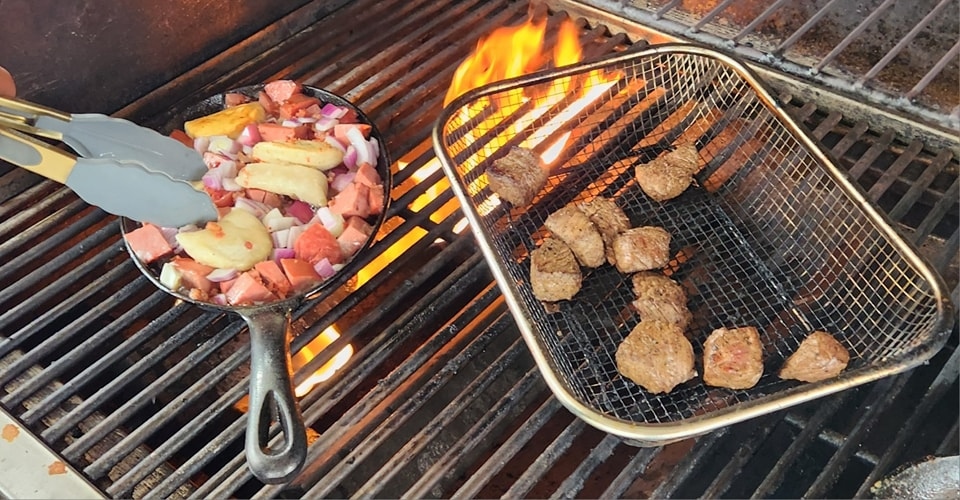 Steak bites grilling in a basket beside a cast-iron pan of potatoes, sausage, and onions over open flames on the Van’s Grill BBQ.